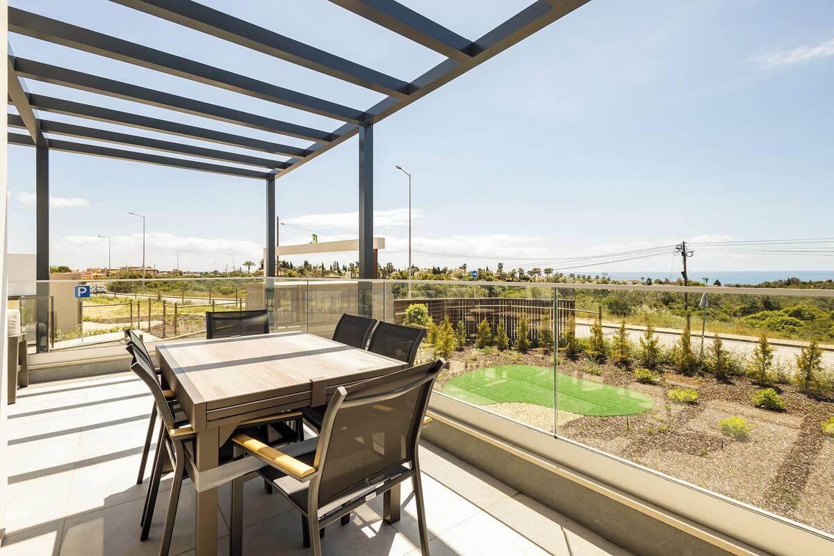 Uma varanda com mesa e cadeiras, vista para uma estrada e vegetação, e um céu claro.