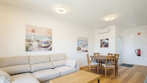 Ein Wohnzimmer mit beiger Couch, Holztisch und einem Essbereich mit Holzsesseln und einem Tisch. An den Wänden befinden sich Gemälde, die Küstenlandschaften zeigen.