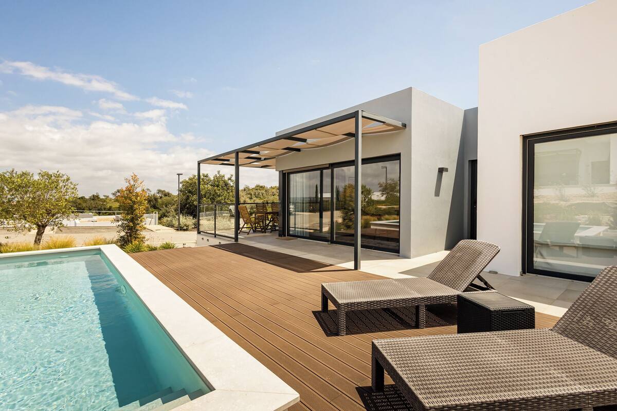 Uma área de piscina moderna ao ar livre, com deck de madeira, espreguiçadeiras e um terraço envidraçado.