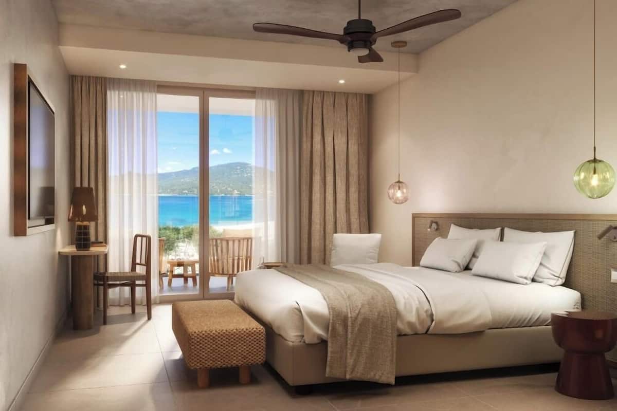 A modern hotel room with a large bed, a desk with a chair, a small table, and a view of the sea through a sliding door.
