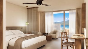 A modern hotel room with a large bed, a ceiling fan, a view of the sea, a small table with books, and a chair. - Casa C (Propriano)