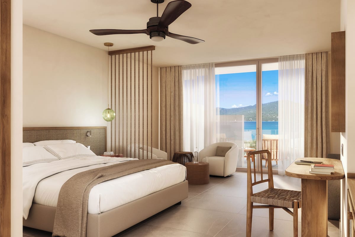 A modern hotel room with a large bed, a ceiling fan, a view of the sea, a small table with books, and a chair.