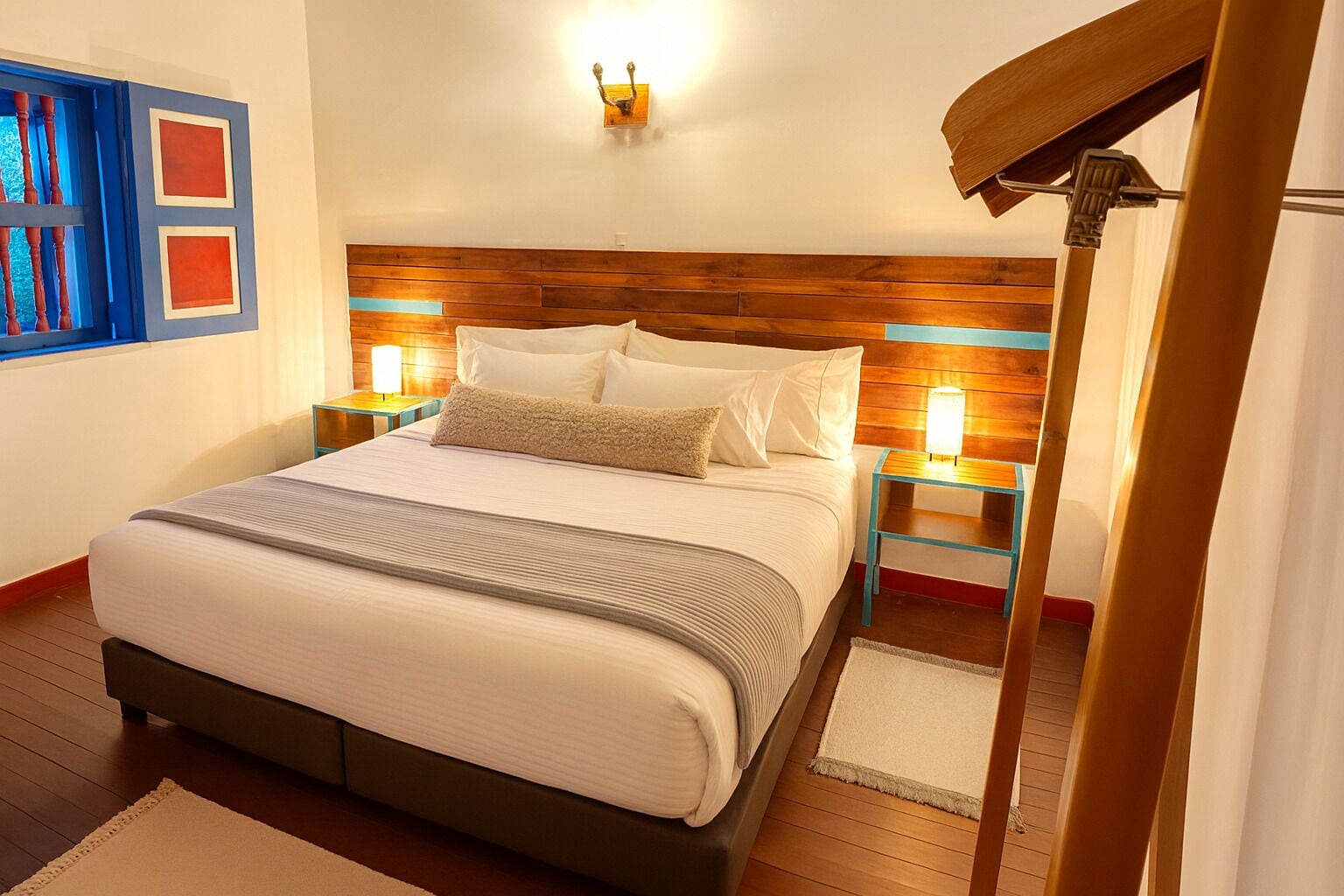 Una cama bien hecha con cabecera de madera, dos mesitas de noche con lámparas, un ventanal con marcos azules y suelo de madera.
