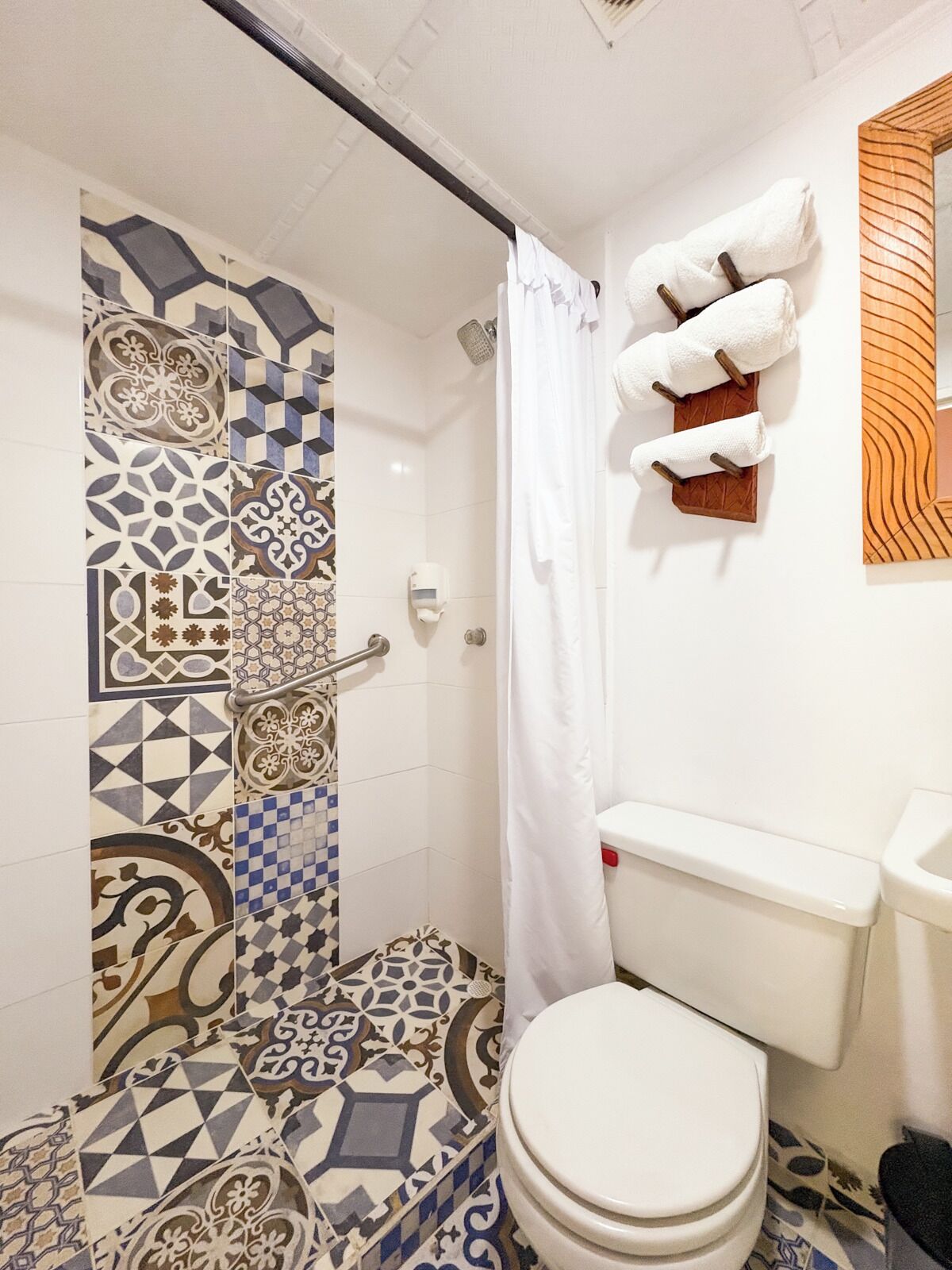 A bathroom with patterned tiles, a white toilet, a shower with a white curtain, and towels on a wooden rack.