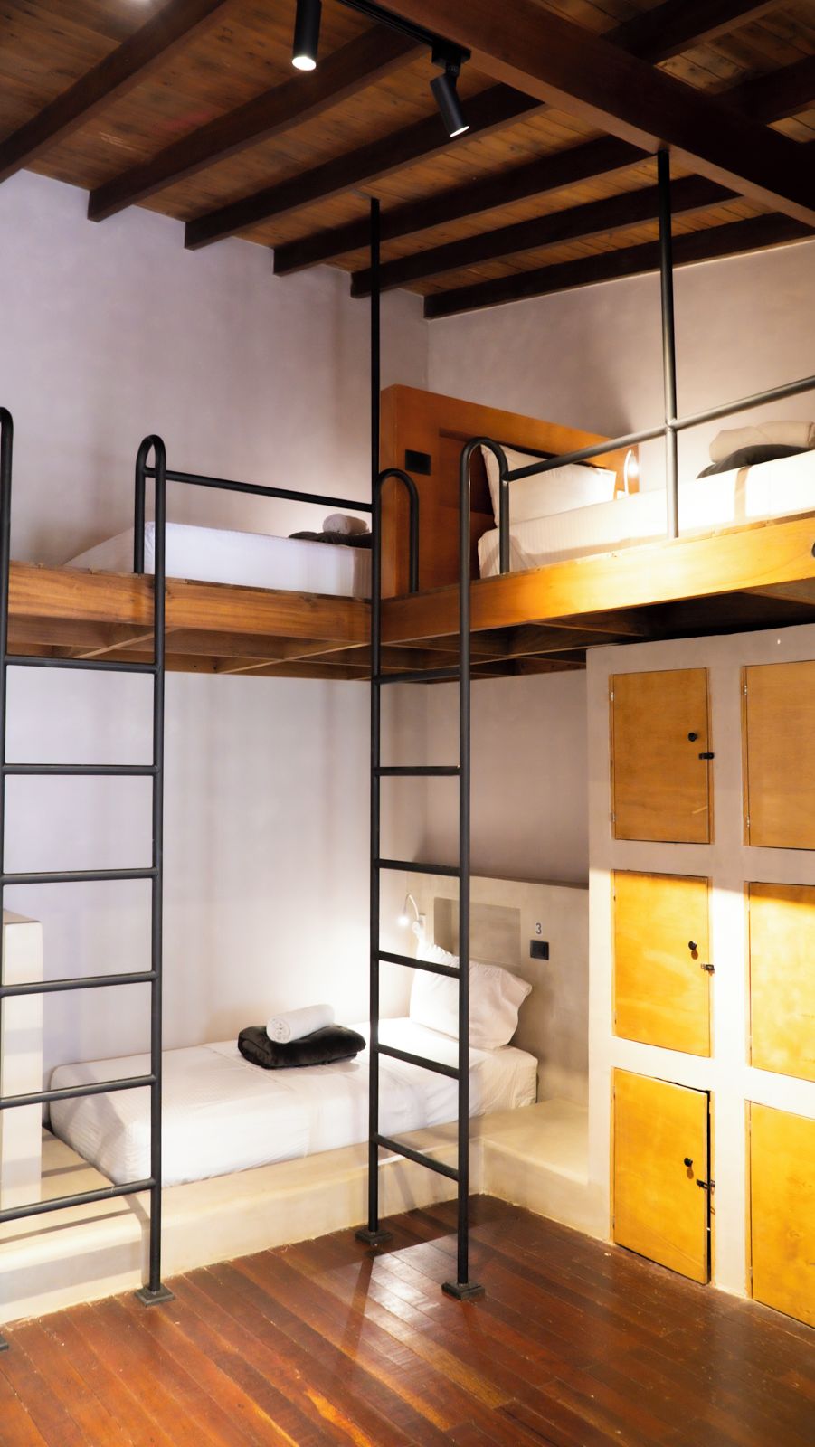 Quarto com beliche, teto de madeira e escada de metal, uma pequena mesa de cabeceira com abajur e guarda-roupa de madeira com três portas.