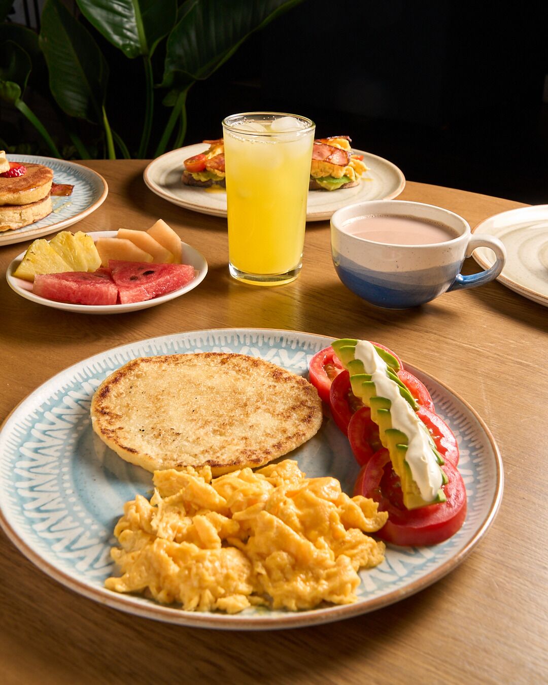 Ein Frühstückstisch mit Rührei, Toast und einem Tomatensalat mit Sahne. Außerdem gibt es Pfannkuchen und ein Glas Orangensaft.
