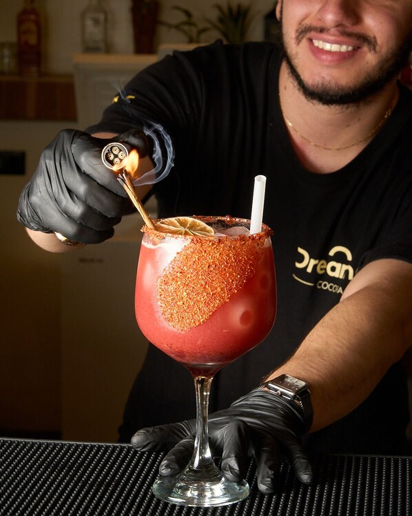 Una persona con guantes y camiseta negros, con la inscripción 'dream' y 'cocoa', prepara un cóctel con una copa en llamas.