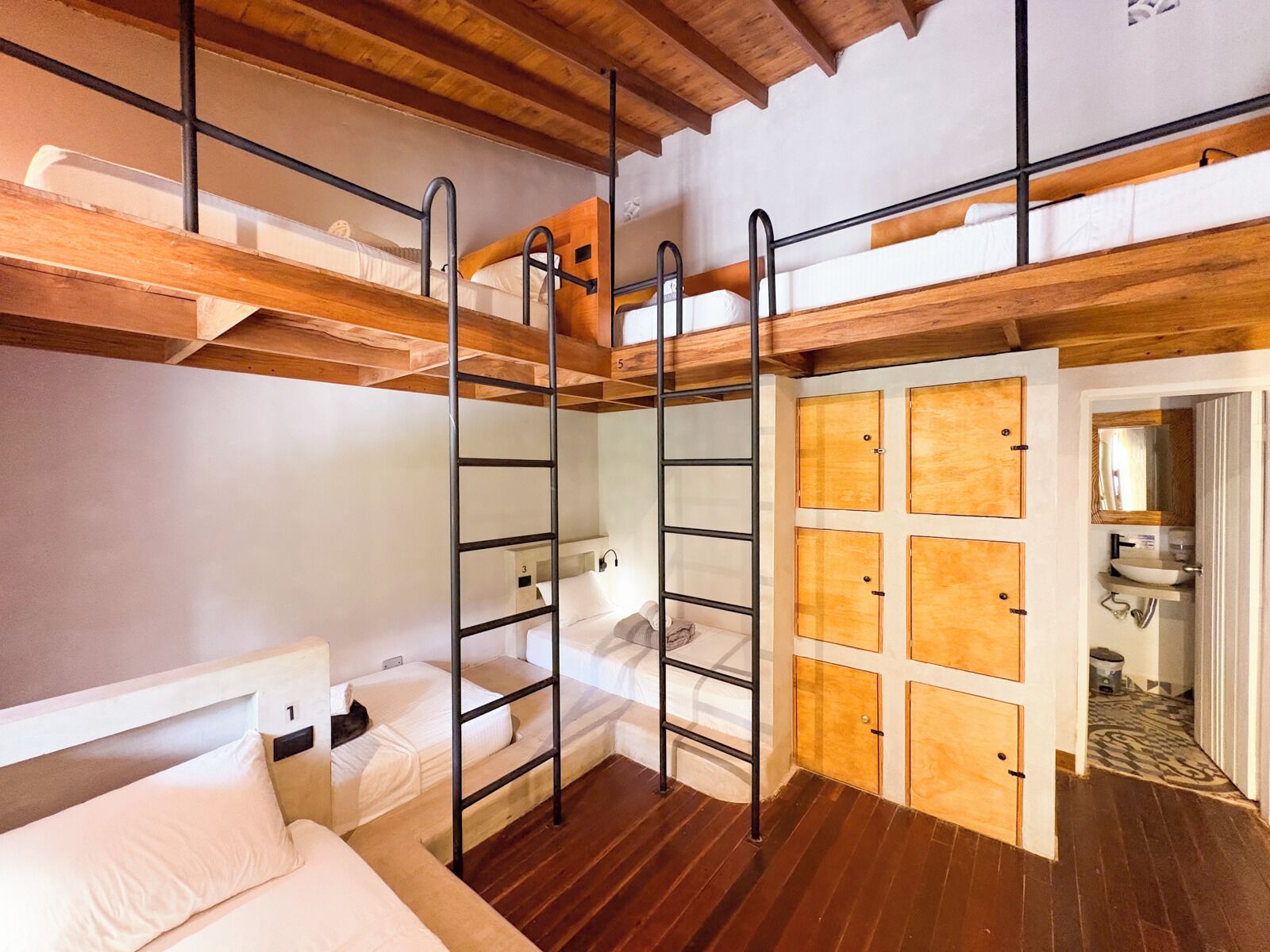 A bunk bed room with a wooden ceiling, a ladder, and a bathroom with a sink and mirror.