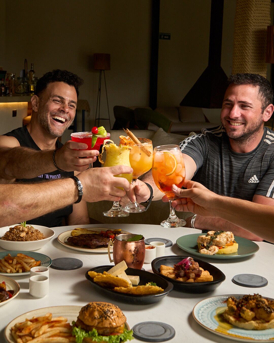 Two people toasting with drinks at a table with various dishes and a copper mug.