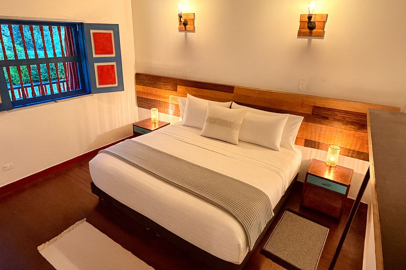 A neatly made bed with white linens and a wooden headboard, two bedside lamps, and a window with a view of greenery.