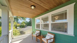 A covered porch with two wooden chairs and a small table, a wooden ceiling, and a green wall with large windows.