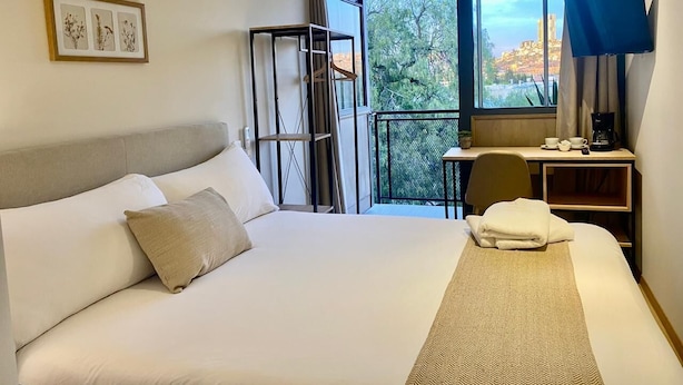 A hotel room with a large bed, a desk with a chair, a television, and a balcony with a view of the city. - Casa Marina (Querétaro)