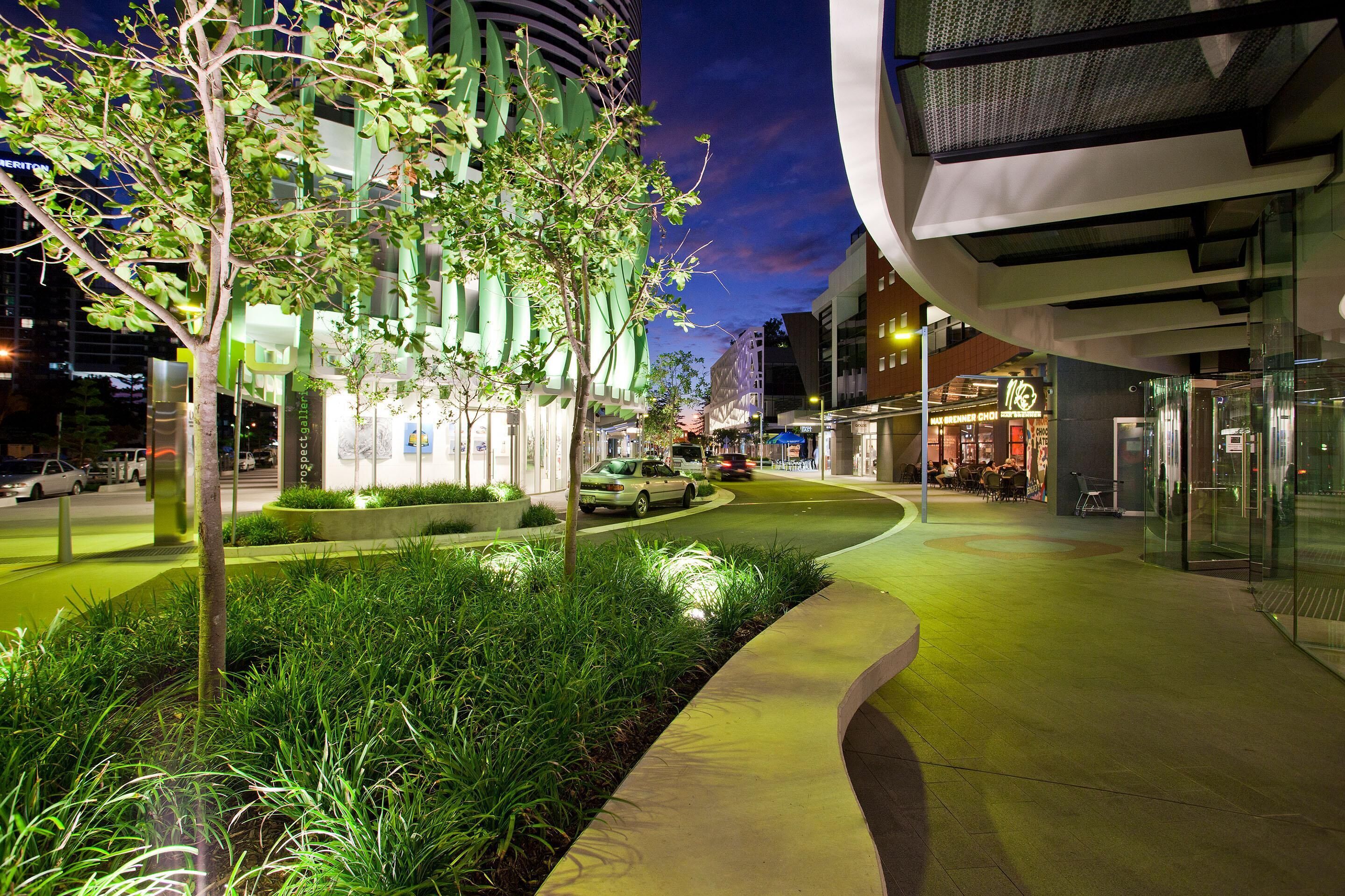 A modern urban street at night with illuminated greenery, a curved sidewalk, and contemporary buildings.