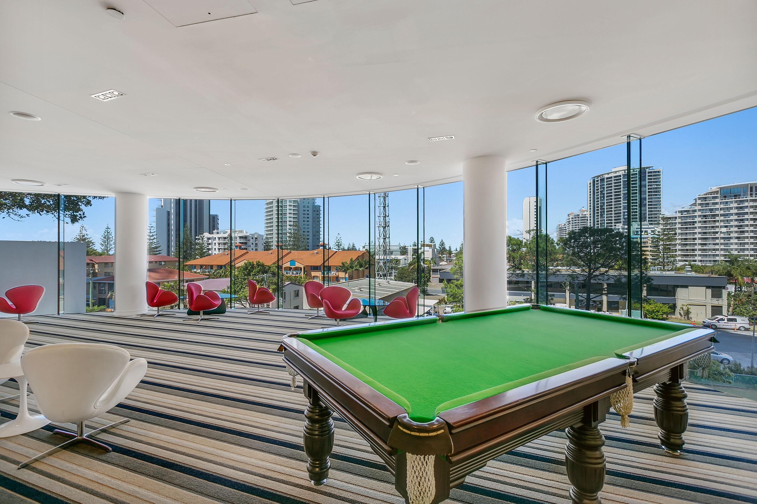 Een pooltafel in een moderne ruimte met grote ramen, rode stoelen en uitzicht op de stad.