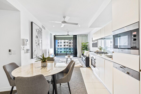 A modern kitchen with a built-in oven and microwave, a dining area with a marble table and chairs, and a view of a residential building through the window. - Cozy 1 Bedroom + Study Unit in Broadbeach - Q Stay (Broadbeach)
