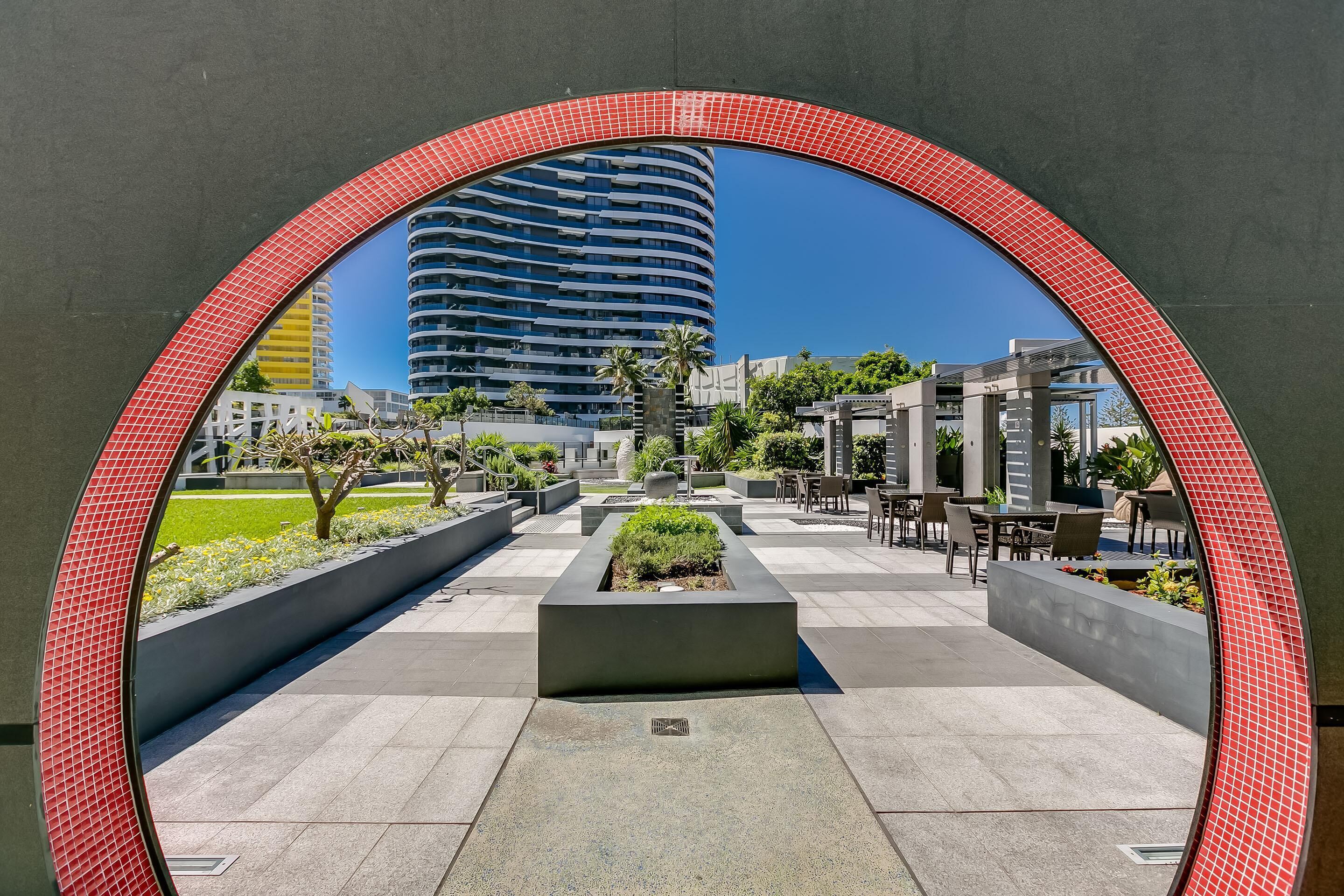 Un espace extérieur moderne doté d’une arche aux tuiles rouges incurvées, d’un jardinier central agrémenté de verdure et d’un coin salon avec des tables et des chaises.