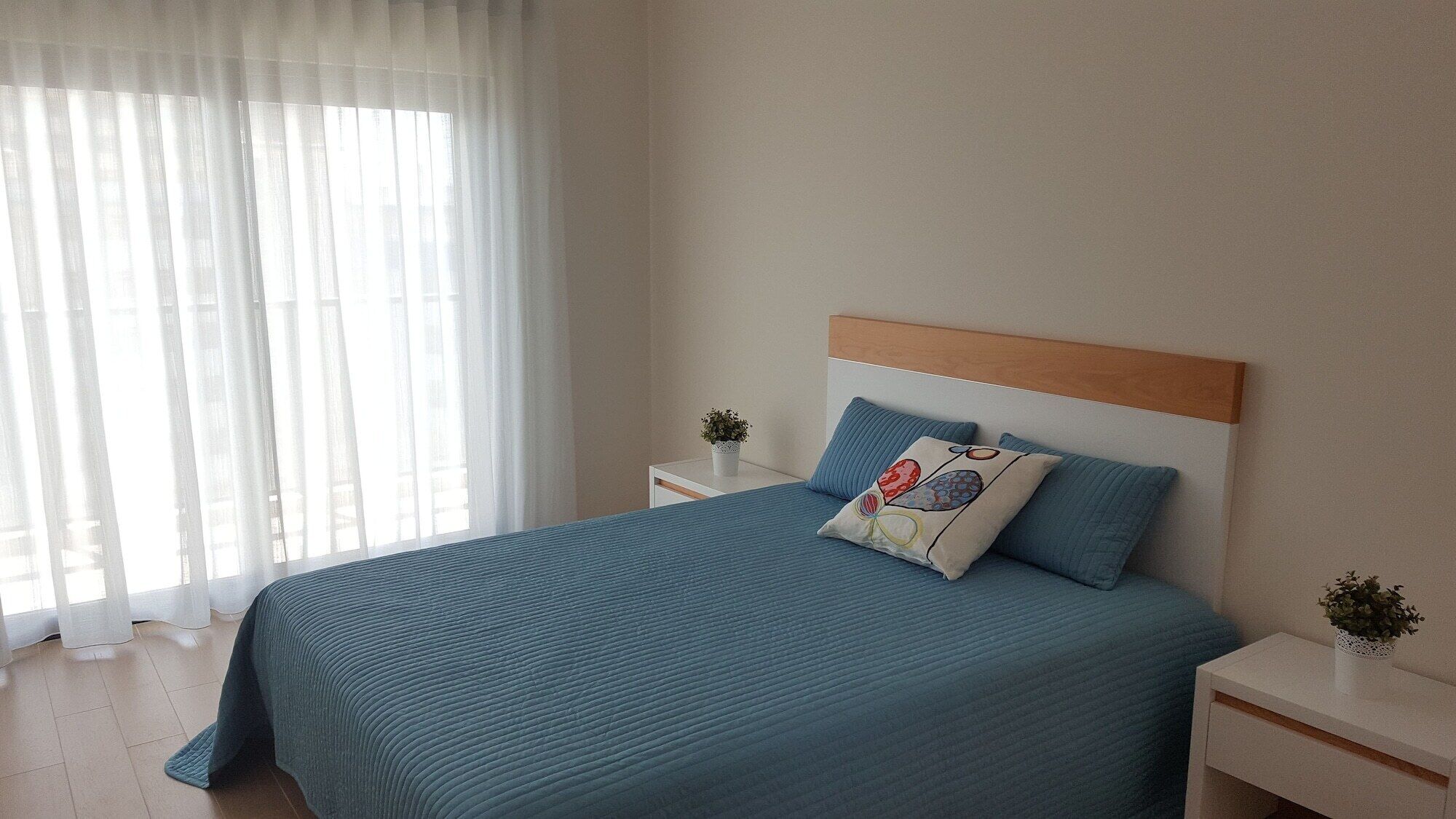 Ein Schlafzimmer mit einem Bett, einem Nachttisch und einem Fenster mit transparenten Vorhängen.