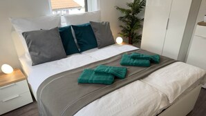 Un lit double avec une tête de lit grise, du linge de lit blanc et des serviettes turquoise. Il y a une table de chevet avec une lampe, une plante en pot et une armoire.