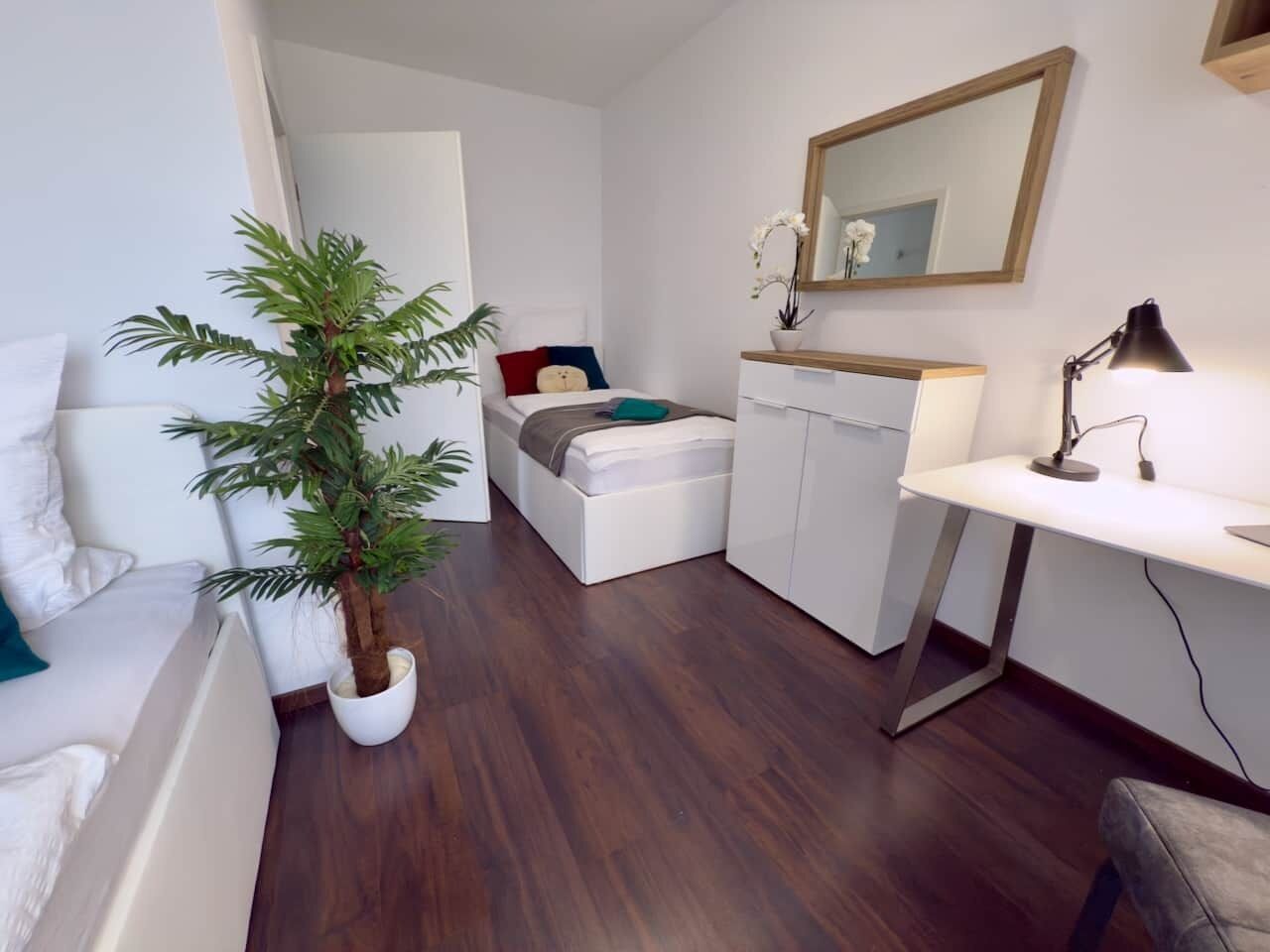 Ein gemütlicher Raum mit Bett, Schreibtisch, Stuhl, einem Topf mit Pflanze und einem Spiegel an der Wand.