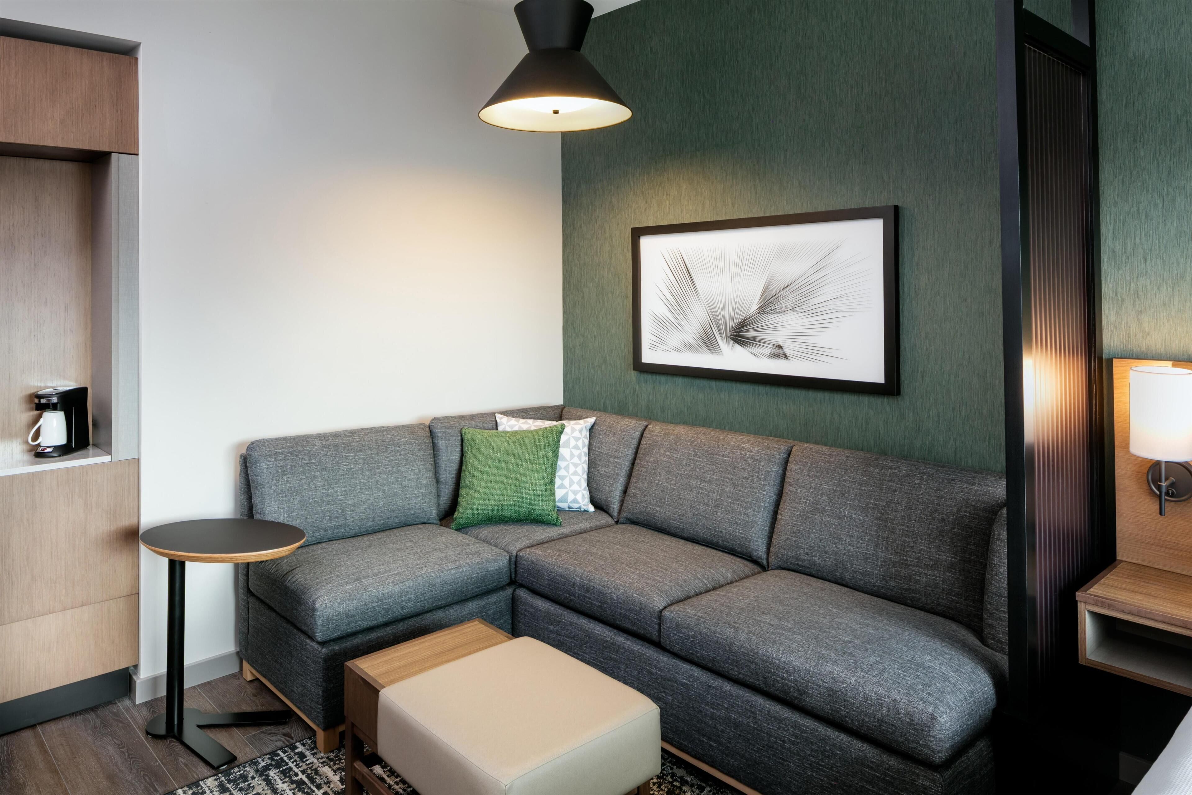 A modern living room with a grey sectional sofa, a small round table, a coffee machine, and a framed abstract art piece on the wall.