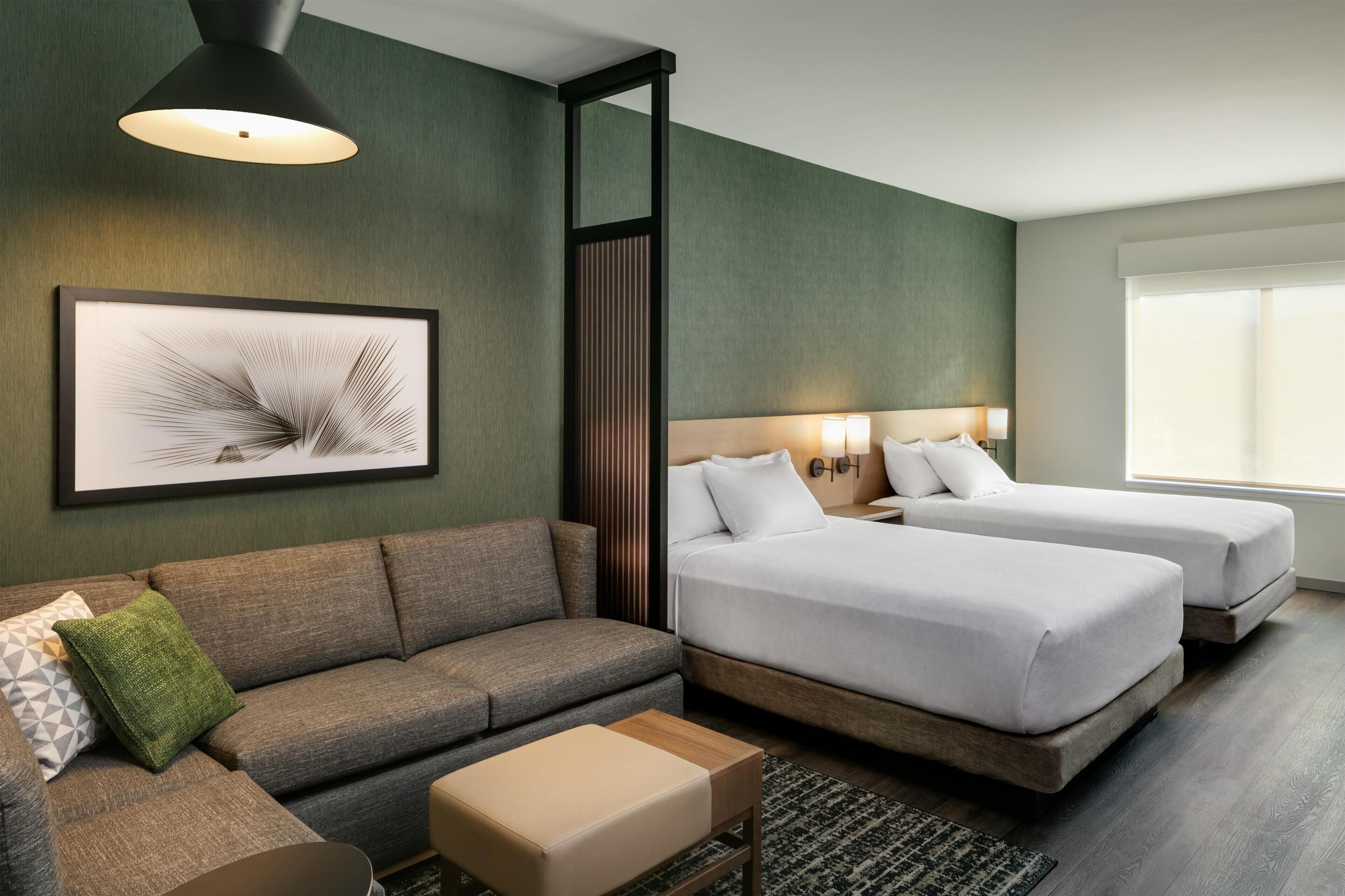 Una habitación de hotel con sofá, dos camas, una obra de arte enmarco y una ventana.