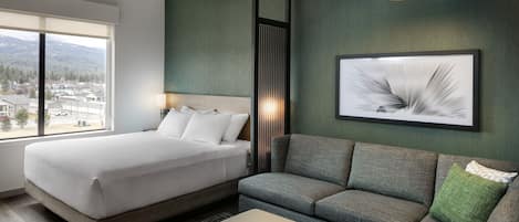 A modern hotel room with a large bed, a sofa, and a small table. There is a window with a view of buildings and mountains.