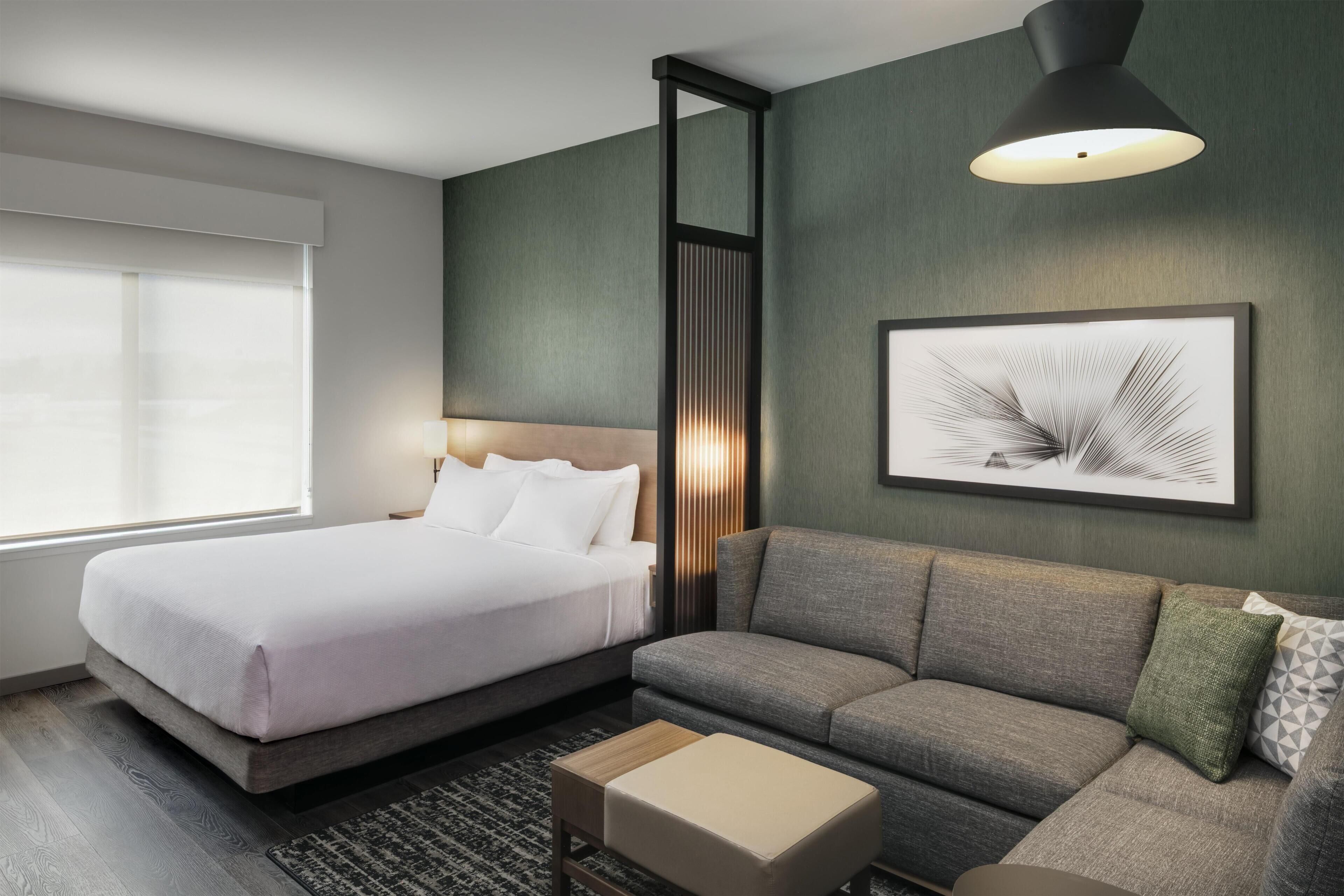 A modern hotel room with a bed, a sofa, a small table, and a framed artwork on the wall.