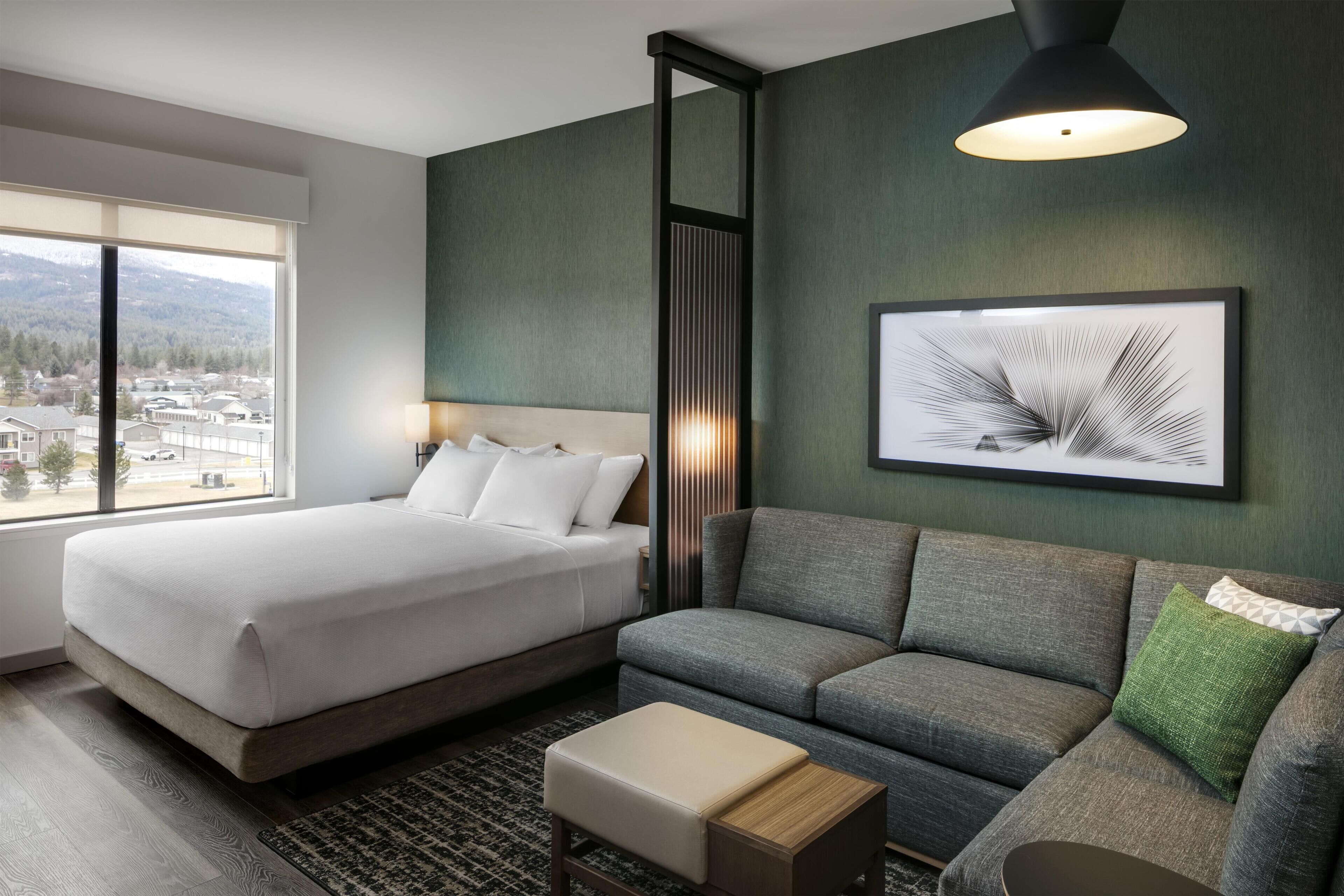 Ett hotellrum med en säng, en soffa, ett litet bord, en inramad tavla och ett fönster med utsikt över byggnader och berg.