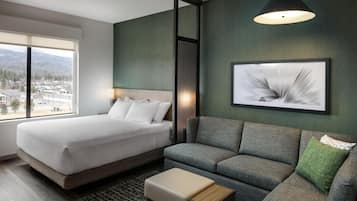 A hotel room with a bed, a sofa, a small table, a framed picture, and a window with a view of buildings and mountains.