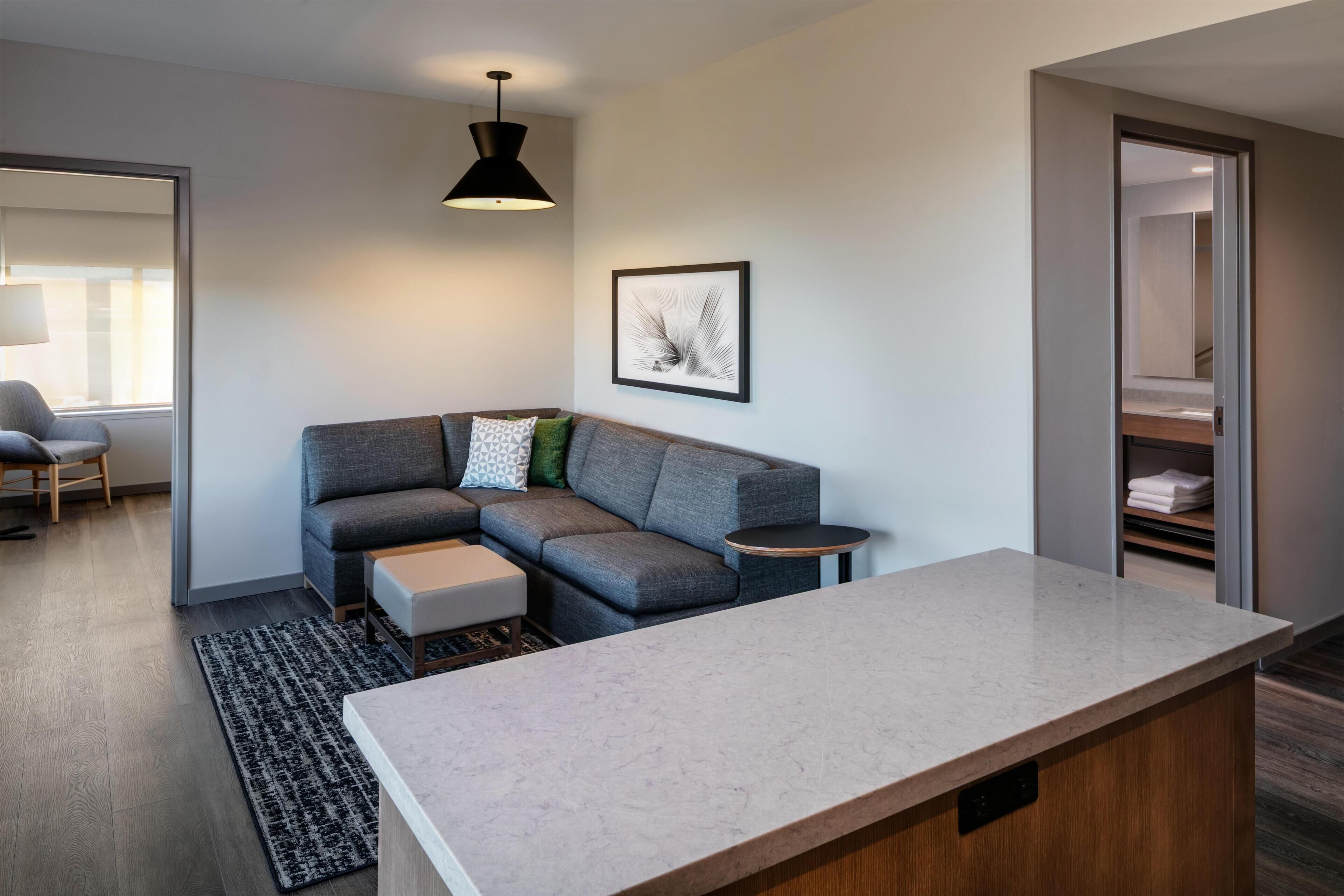 A modern living room with a grey sectional sofa, a small round table, a marble countertop, and a black pendant light.