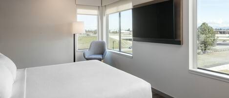 Habitación de hotel con una cama, una silla, un televisor montado en la pared y grandes ventanales con vistas al exterior.