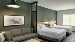 Et moderne hotelværelse med en grå sofa, to enkeltsenge, et lille bord og et indrammet abstrakt kunstværk på væggen.