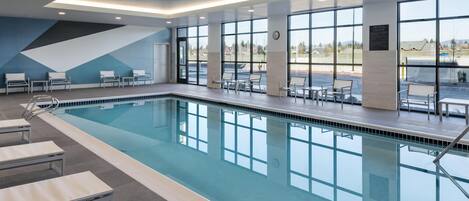 Una piscina interior moderna con paredes de vidrio transparente, zonas de descanso y vistas al estacionamiento.