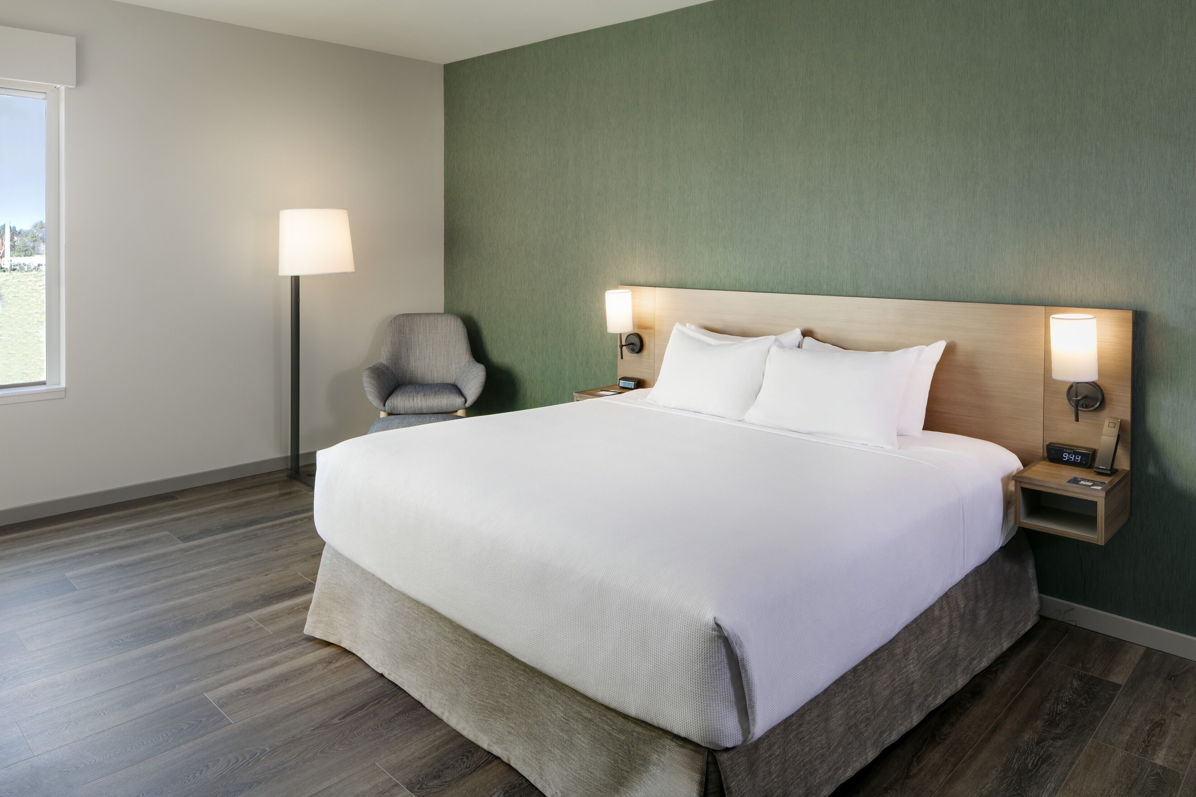 A neatly made bed with a wooden headboard, a bedside table with a lamp, and a chair in a room with green walls and wooden flooring.