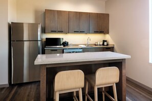 A modern kitchen with a stainless steel refrigerator, a microwave, a coffee maker, and wooden cabinets. There is a large island with two bar stools. - Hyatt Place Post Falls/Coeur D'Alene (Post Falls)