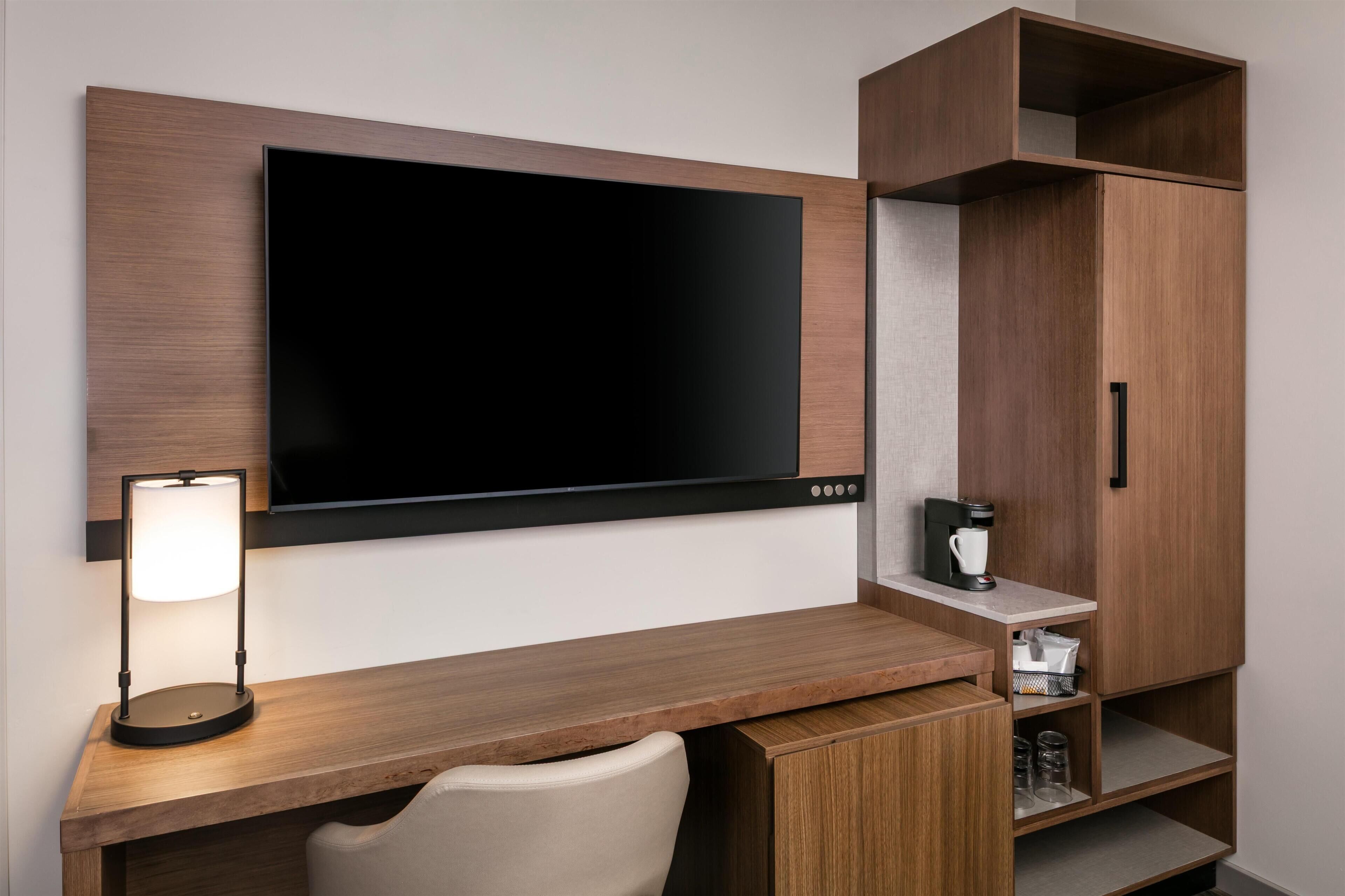 Una habitación de hotel moderna con una televisión de pantalla plana montada en la pared, un escritorio con lámpara, una silla y armarios empotrados.