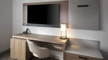 A modern hotel room with a wooden desk, a flat-screen TV, a desk lamp, a chair, and a small drawer.