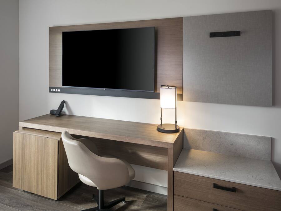 A modern hotel room with a wooden desk, a flat-screen TV, a desk lamp, a chair, and a small drawer.
