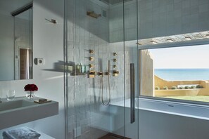 A modern bathroom with a glass shower enclosure, a large mirror, and a view of the sea. - Vila Joya (Albufeira)