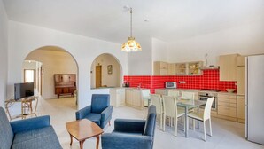 A spacious living area with a TV, blue armchairs, a wooden coffee table, and a dining table with chairs. A kitchen with red tiles and modern appliances is visible in the background.