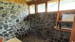 Un baño con paredes de piedra, una bañera, zona de ducha y un estante de madera con toallas.