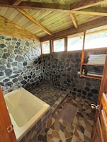 Un baño con paredes de piedra, una bañera, zona de ducha y un estante de madera con toallas.