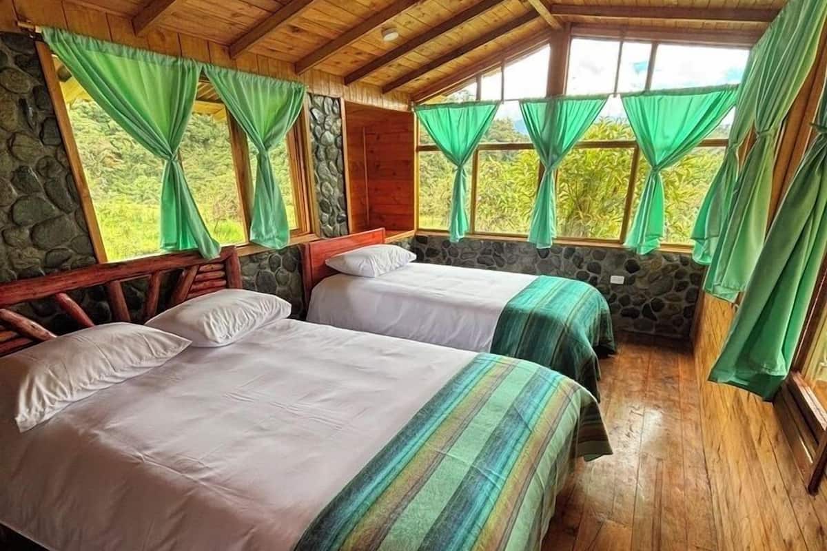 Una cabaña de madera con dos camas, paredes de piedra y ventanas amplias con cortinas verdes.