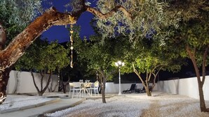 Área de terraço ao ar livre com mesa e cadeiras, cercada por árvores e uma parede branca.