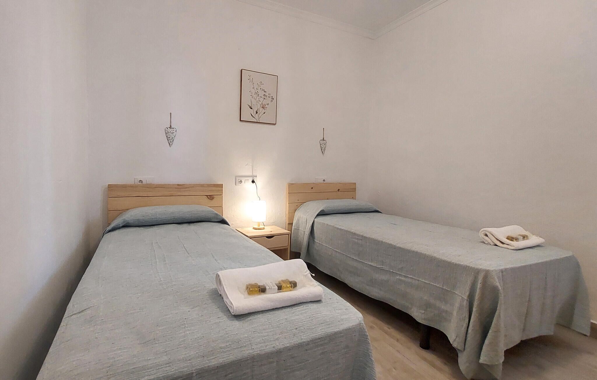 A room with two single beds, each with a wooden headboard and a light blue bedspread. There is a nightstand with a lamp on one side and a folded towel on the other. A framed picture hangs on the wall above the beds.