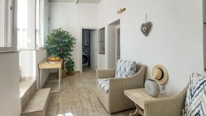 Uma sala de estar clara e arejada, com móveis de vime, uma planta em vaso e vista para a cozinha.