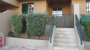 Un bâtiment doté d’un escalier menant à une entrée fermée par un portail, entouré de verdure et bordé d’un trottoir dallé.