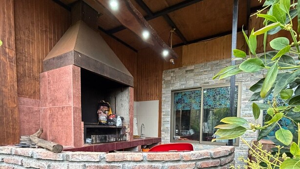 A brick-built outdoor kitchen with a chimney, surrounded by green plants and a glass wall. - CASA EL CANELO (El Quisco)