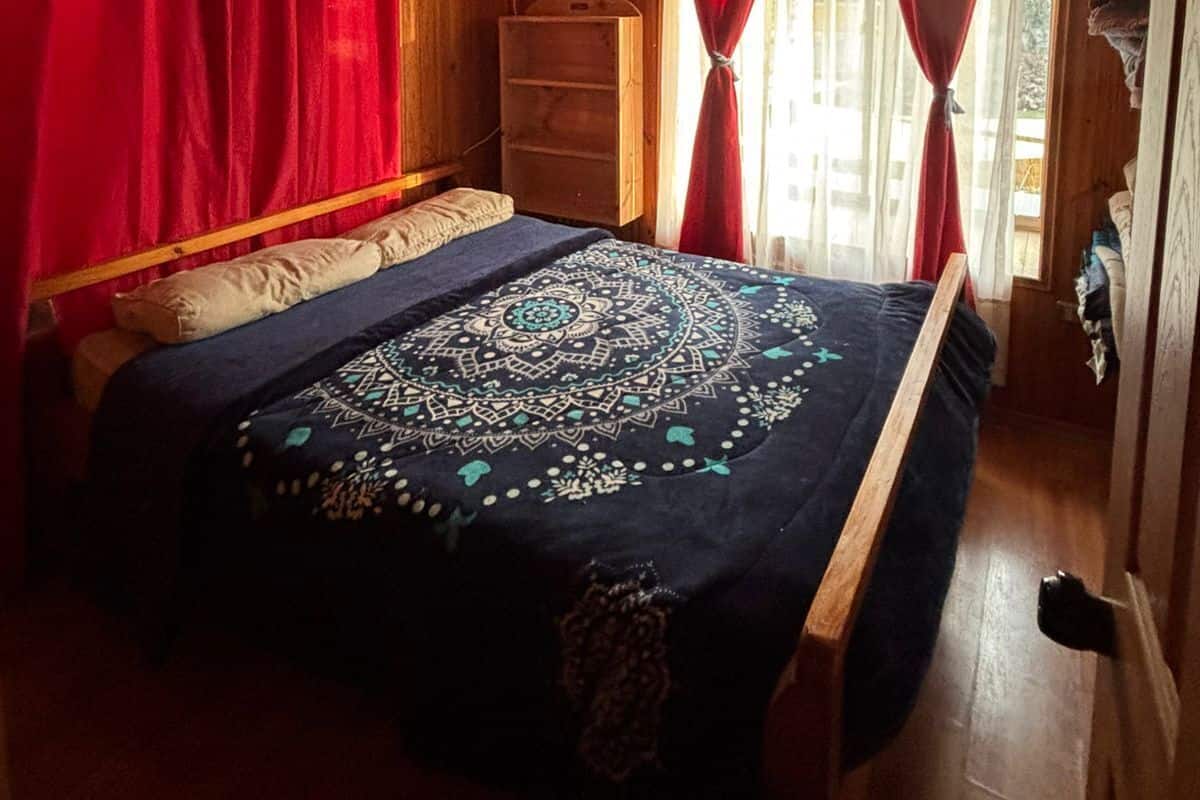 A wooden cabin bedroom with a bed, a dresser, and a window with red curtains.