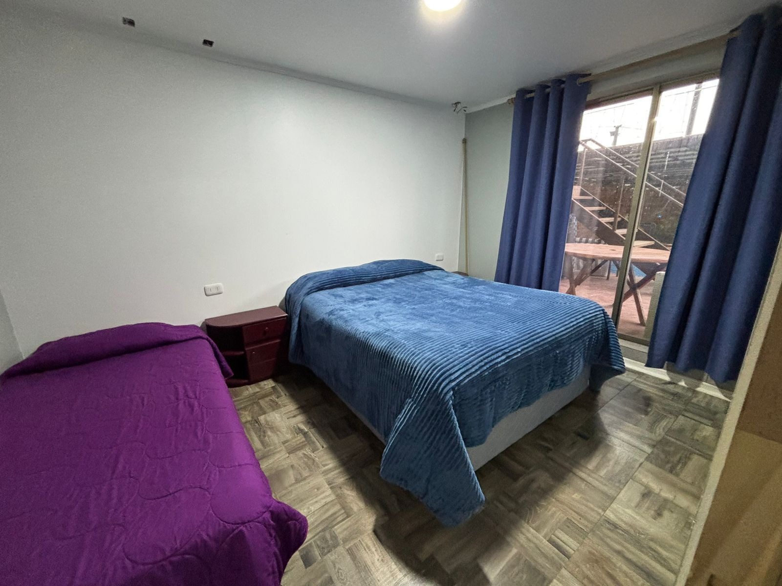 A bedroom with two beds, a wooden floor, a small bedside table, and a window with curtains.
